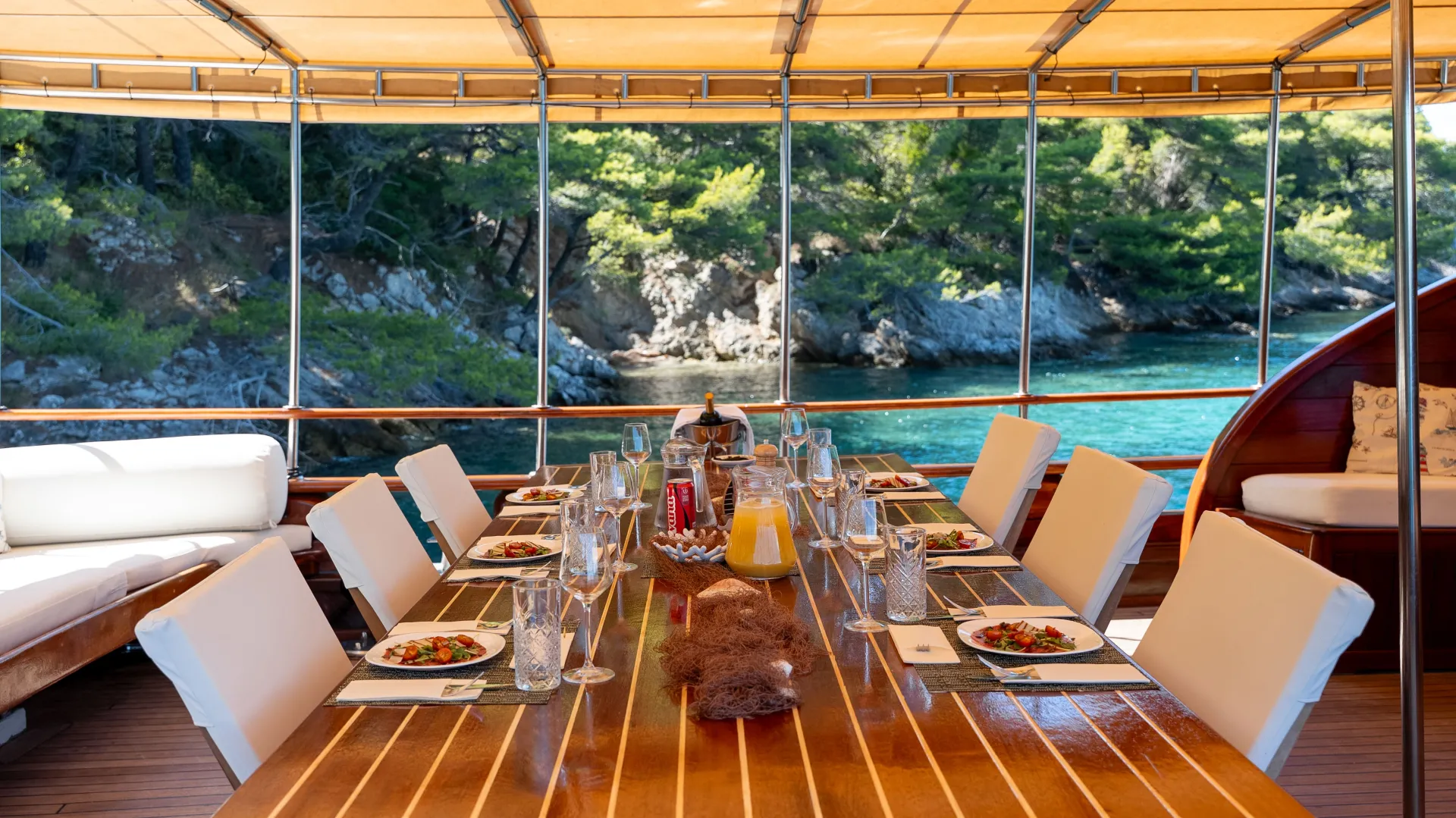 A set dining table with plates of salad, glasses, and drinks on a wooden boat deck, overlooking clear blue water and a lush green rocky shoreline under a canopy.