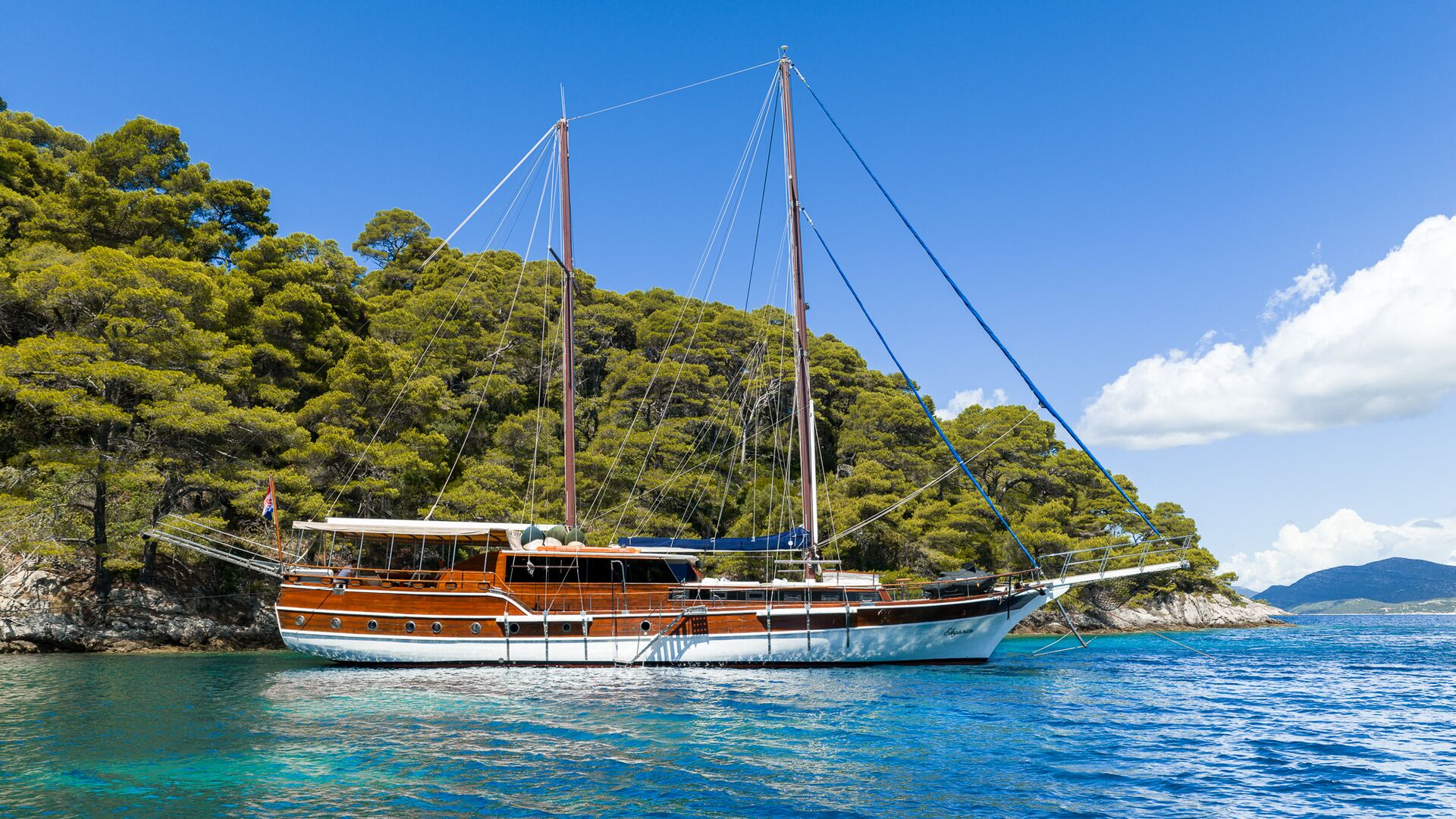 A large wooden sailboat anchored in clear blue water near a lush, tree-covered island under a bright blue sky with a few clouds.