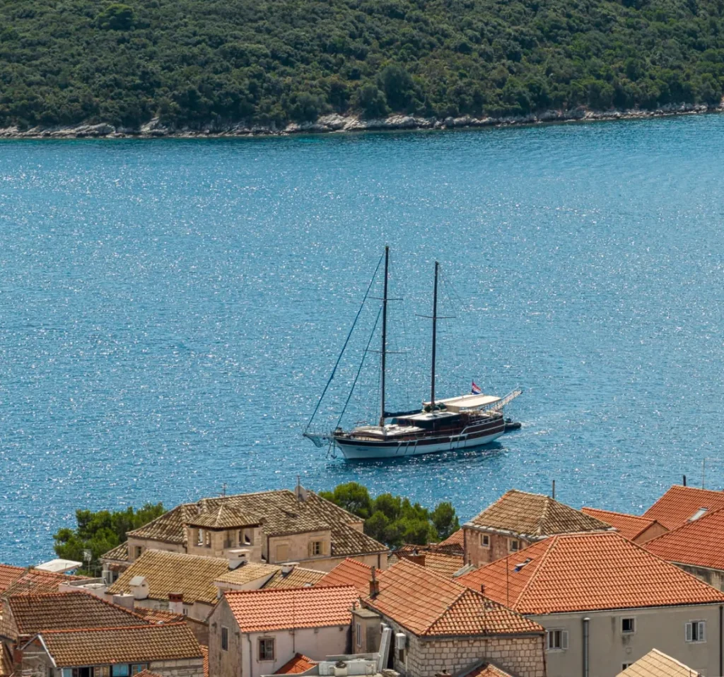 A sailboat floats on blue water near a coastal town with red-tiled rooftops and a stone church tower. Lush green hills line the background across the bay.