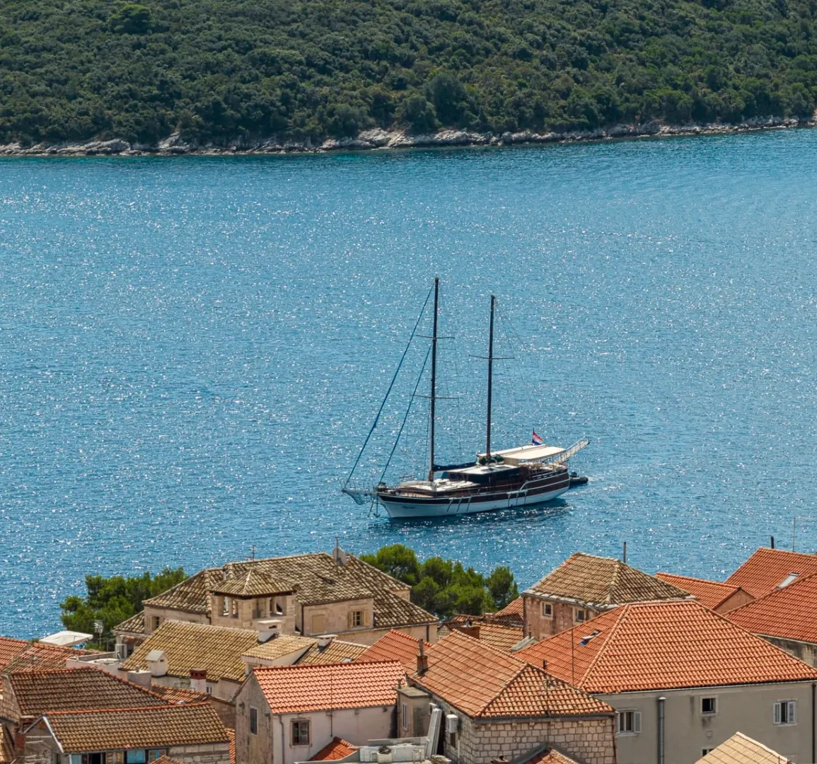 A sailboat floats on blue water near a coastal town with red-tiled rooftops and a stone church tower. Lush green hills line the background across the bay.