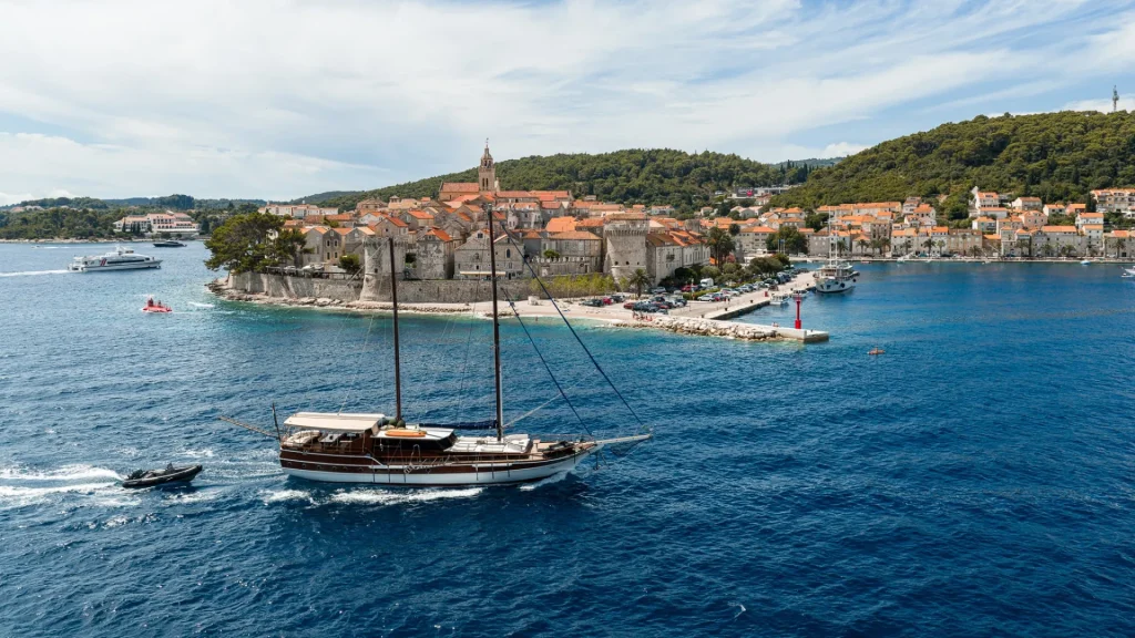 A sailing boat glides on deep blue water near a coastal town with orange-roofed buildings, historic stone walls, and green hills under a partly cloudy sky.