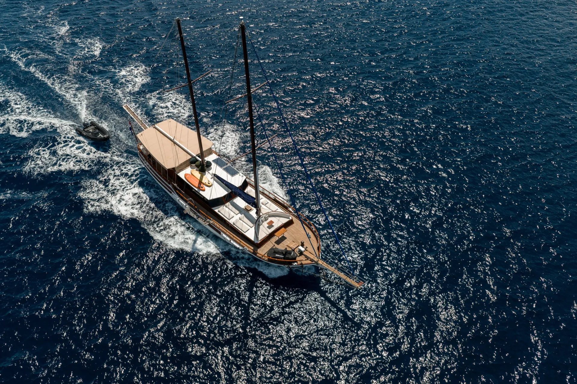 Aerial view of a large sailboat with two masts sailing on deep blue water, leaving a white wake behind. A small motorboat trails closely beside the yacht under clear, sunny skies.
