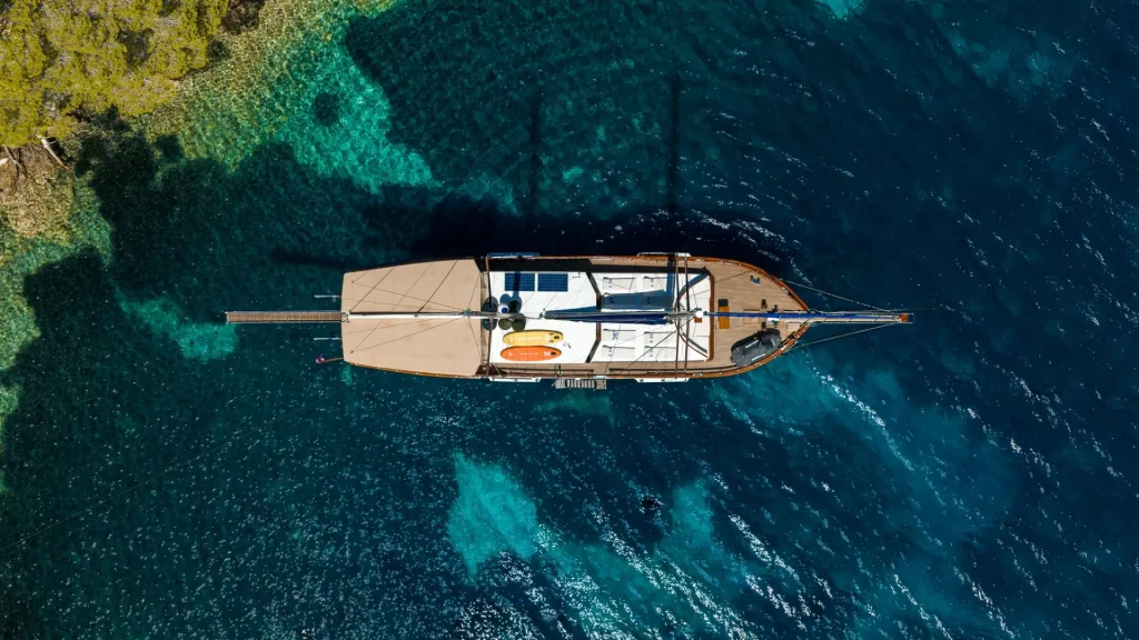 Aerial view of a wooden sailboat anchored in clear blue water near a shoreline with trees and shallow, turquoise areas visible beneath the surface.