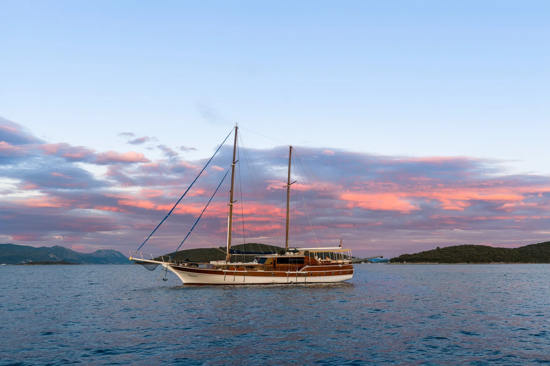 A white sailboat with two masts floats on calm blue water at sunset, with pink clouds and distant green islands in the background.