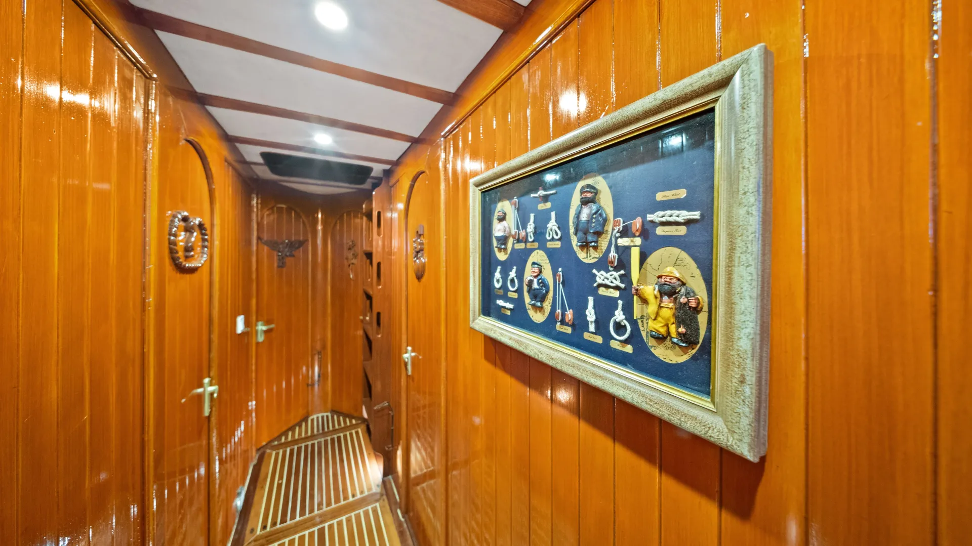 A narrow hallway with glossy wooden walls and ceiling, decorated with framed nautical memorabilia and wooden door accents, leading to several doors and a patterned wooden floor.