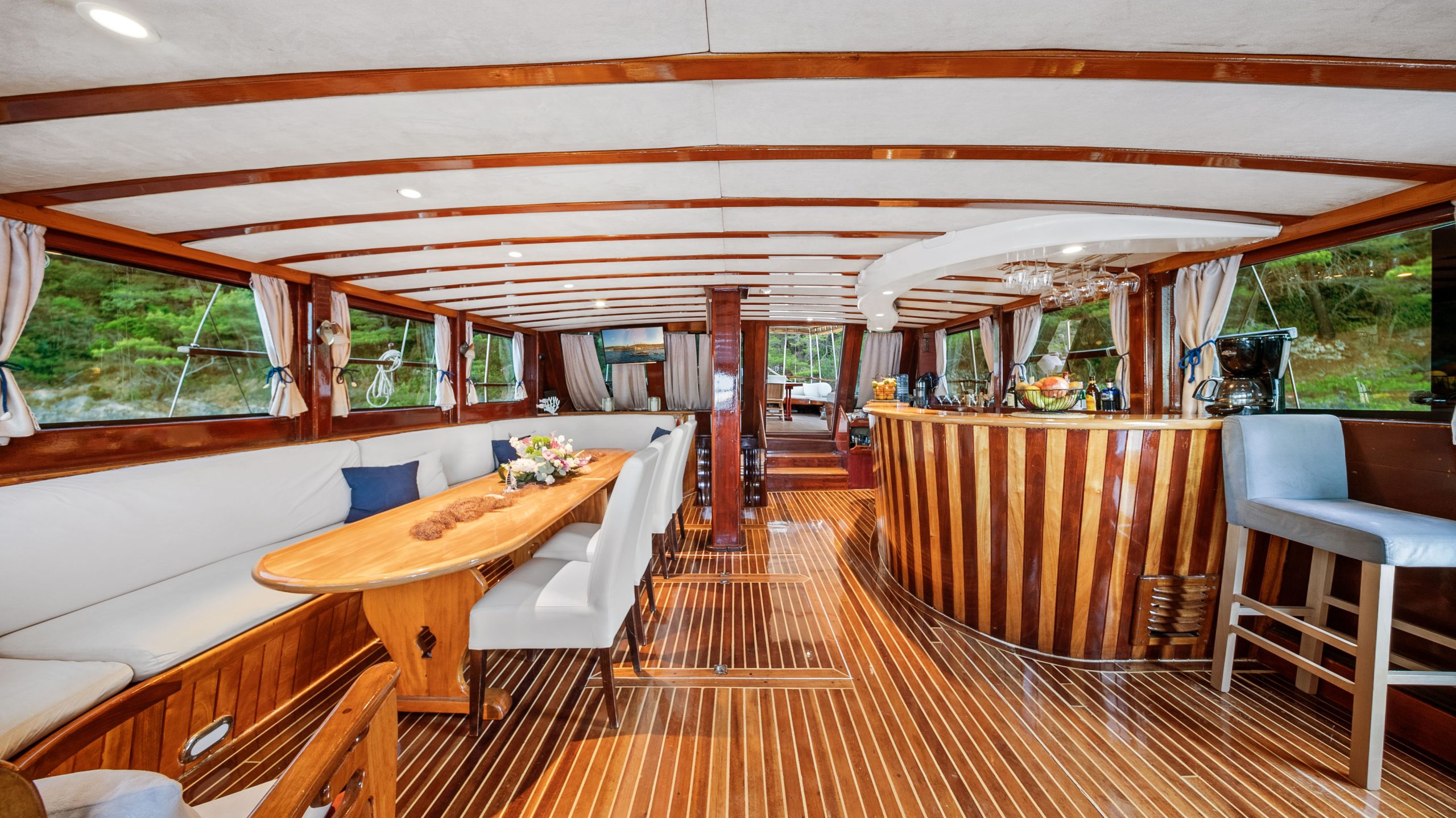 Interior of a luxury yacht with polished wooden floors and ceiling, a curved wooden bar with stools, a long table with seating, and large windows offering views of greenery outside.