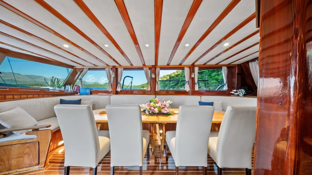 A luxurious yacht dining area featuring a wooden table with a flower arrangement, surrounded by eight white cushioned chairs, large windows with scenic water and greenery views, and polished wood paneling throughout.