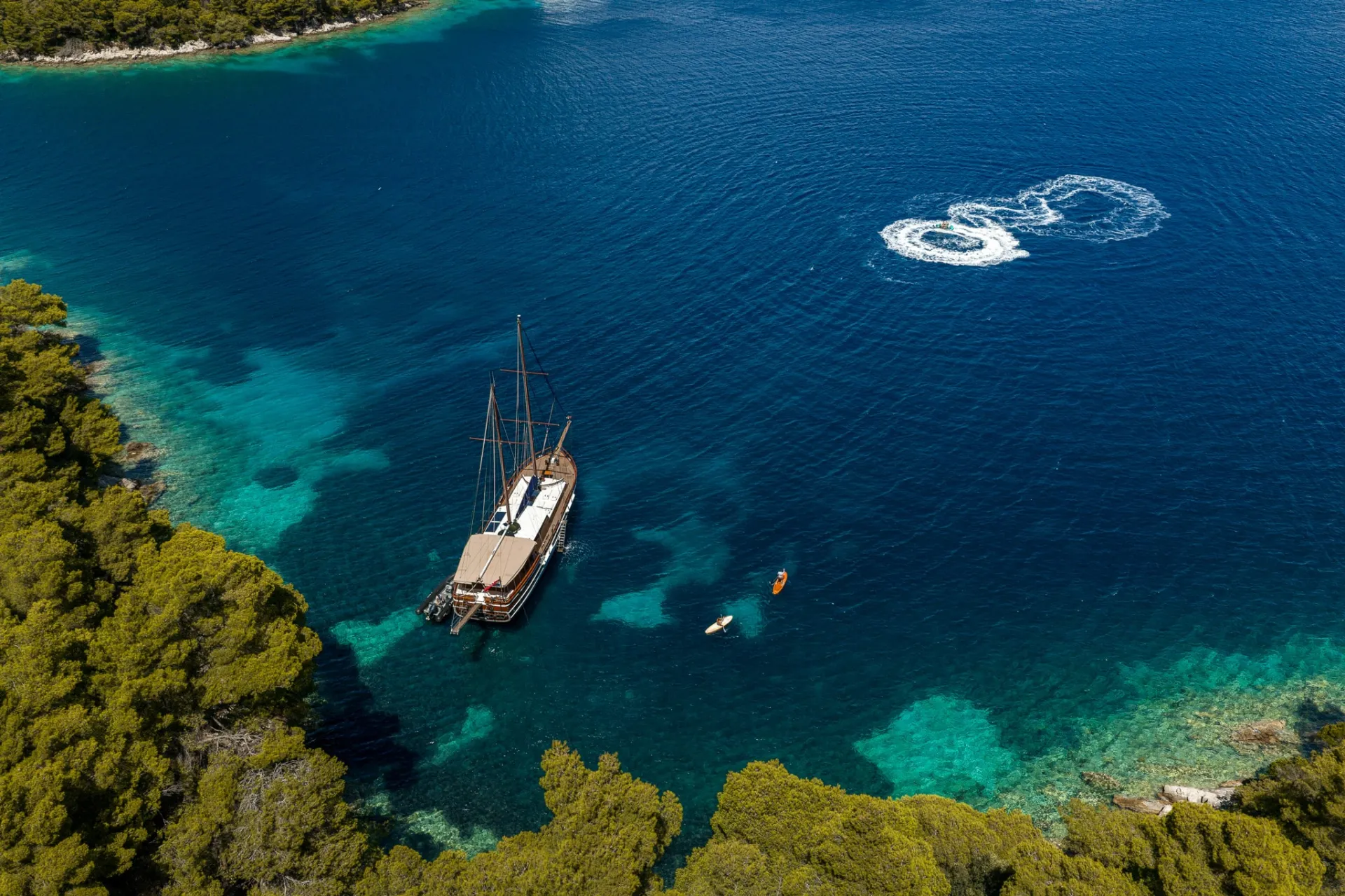 Aerial view of a wooden sailing ship anchored in clear blue water near a forested coastline, with people in kayaks and visible circular patterns in the water nearby.