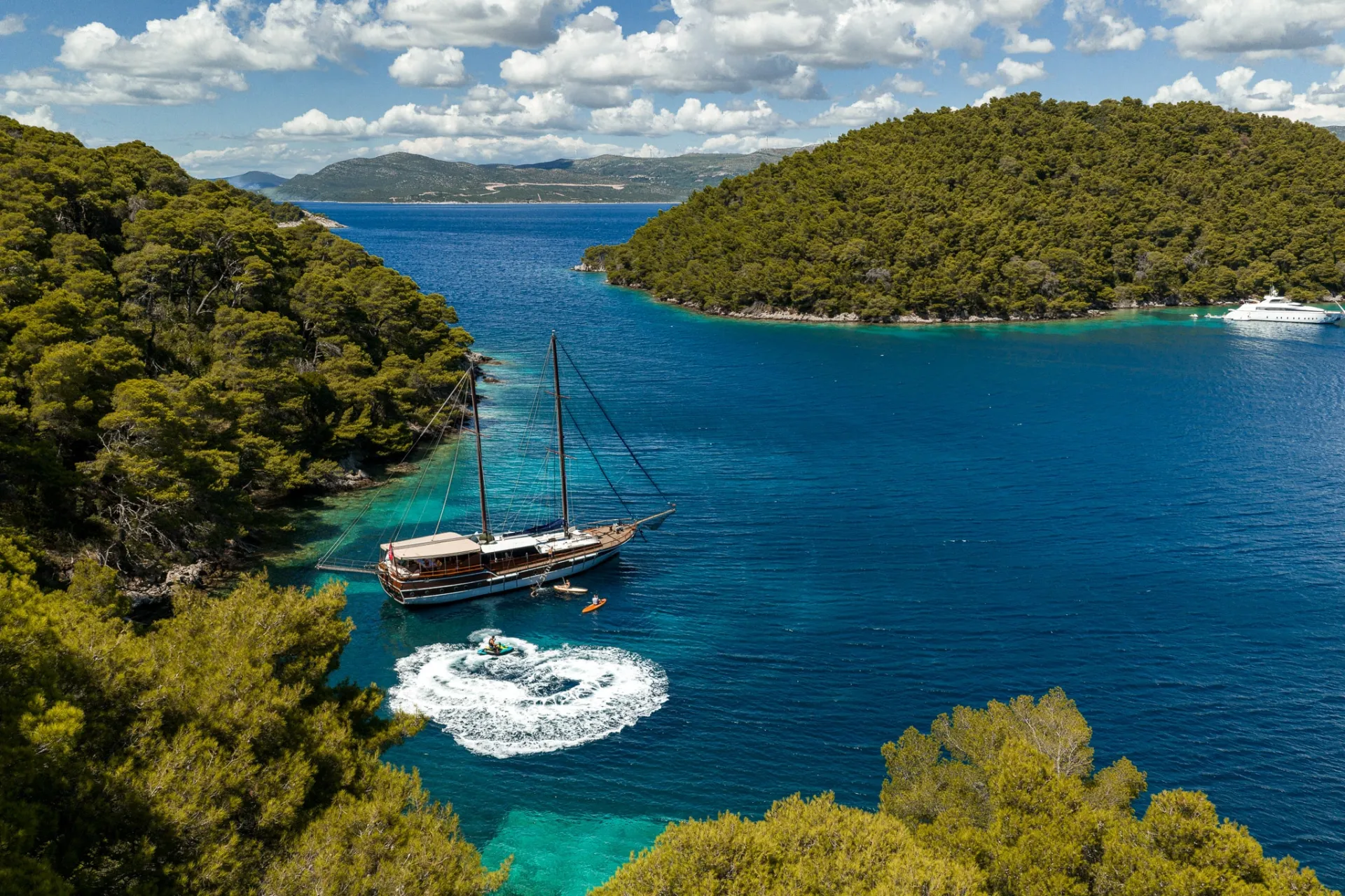 A wooden sailboat and a small motorboat are anchored in a clear blue bay surrounded by lush, green forested hills under a partly cloudy sky. Another yacht is visible in the distance.