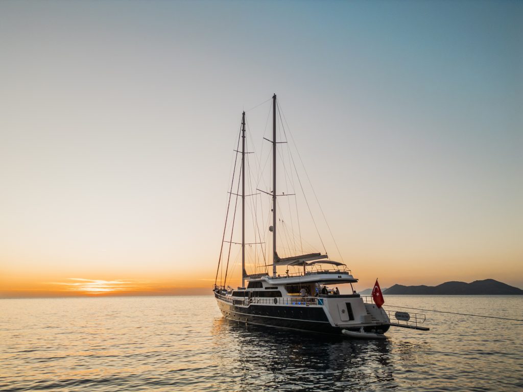 A large sailboat with two masts floats on calm water at sunset, with an orange sky and distant mountains in the background.