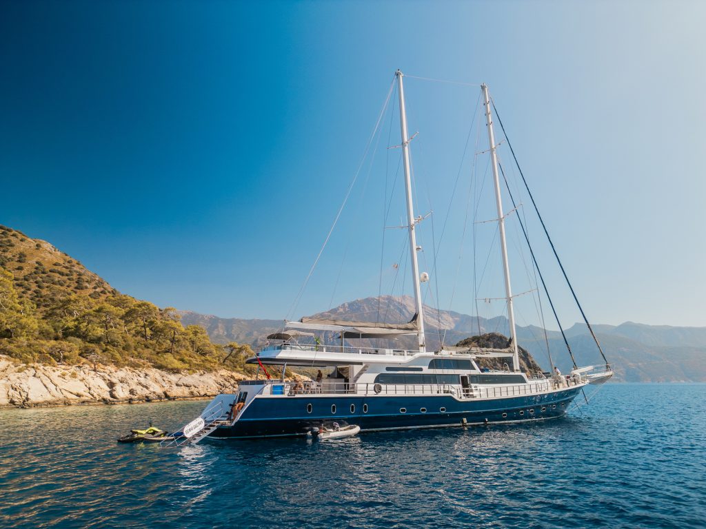 A large, luxurious sailing yacht with two tall masts is anchored near a rocky coastline with green trees, set against clear blue water and a mountainous backdrop under a bright, sunny sky.