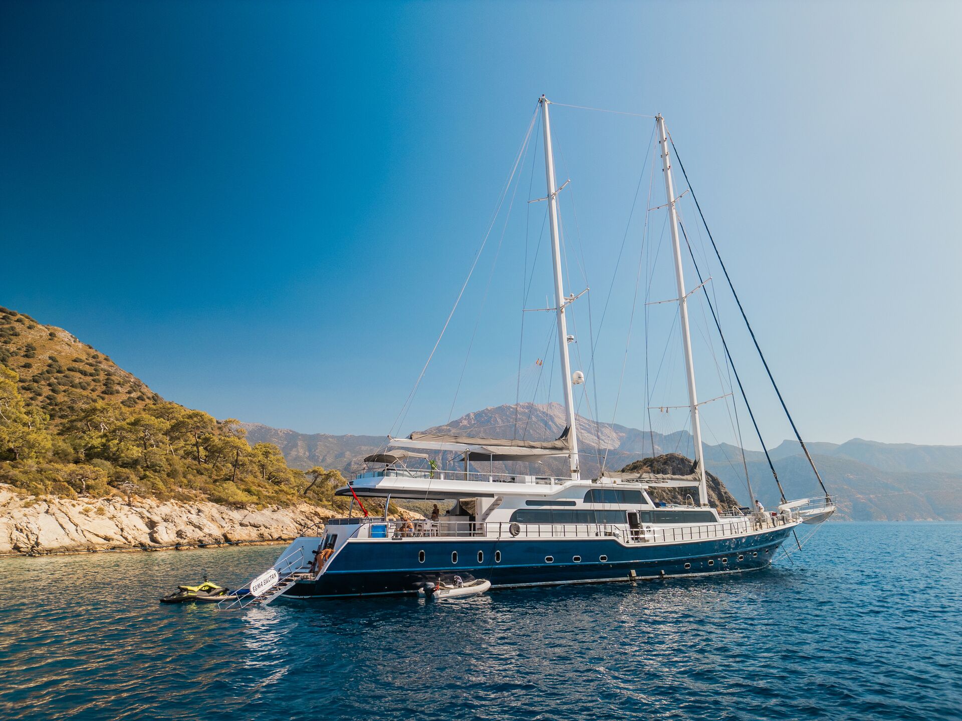 A large, luxurious sailing yacht with two tall masts is anchored near a rocky coastline with green trees, set against clear blue water and a mountainous backdrop under a bright, sunny sky.
