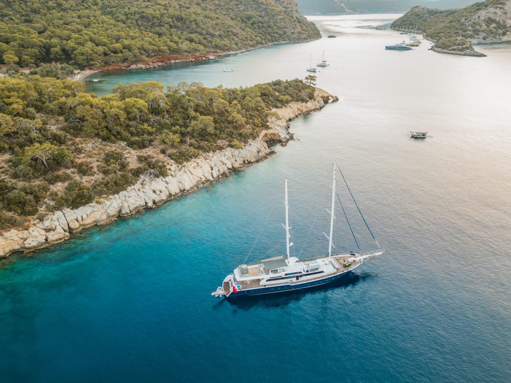 A large sailboat with two masts floats on clear blue water near a rocky, forested coastline with several smaller boats in the distance under a bright sky.