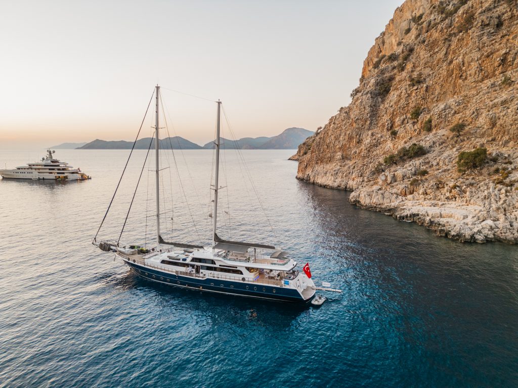 A luxury sailboat with a Turkish flag floats near a rocky coastline at sunset, with another yacht anchored in the distance and mountains visible on the horizon.
