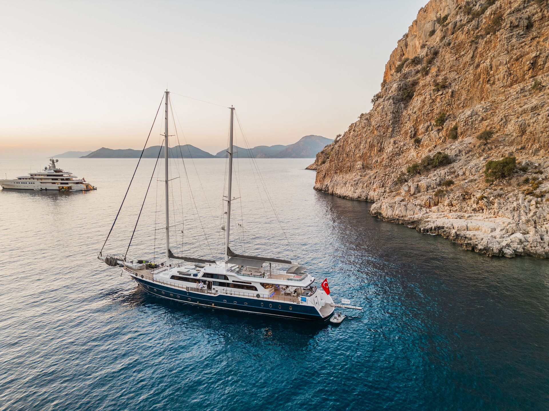 A luxury sailboat with a Turkish flag floats near a rocky coastline at sunset, with another yacht anchored in the distance and mountains visible on the horizon.