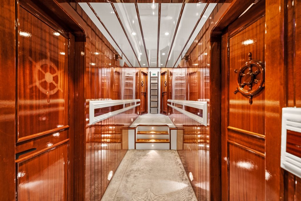 Luxurious hallway with polished wood paneling, nautical wheel designs on doors, soft carpet, mirrored ceiling, and illuminated steps leading to another level, resembling the interior of a yacht or ship.