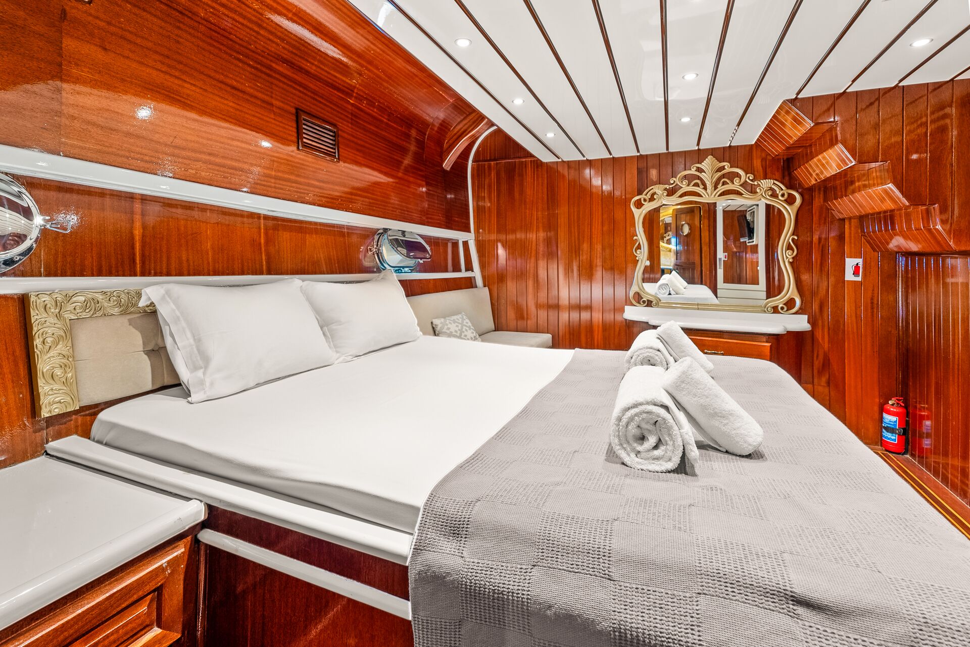 A luxurious cabin with polished wooden walls, a large bed with white linens and neatly rolled towels, a decorative gold-framed mirror above a small vanity, and a fire extinguisher in the corner.