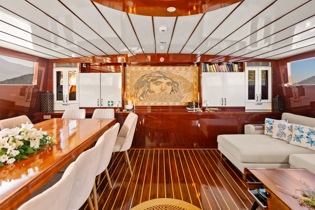 A luxurious yacht interior featuring glossy wood paneling, a dining table with white chairs, a comfortable white sofa with blue cushions, and a mosaic artwork of a face on the back wall. Shelves, windows, and flowers complete the space.