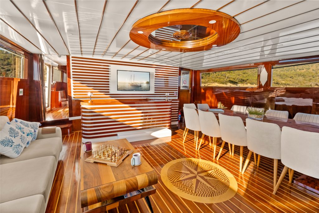 A luxurious yacht interior with wooden floors, a chessboard on a coffee table, cushioned chairs around a dining table, and large windows showing scenic greenery outside. Sunlight streams in, creating a warm, inviting atmosphere.