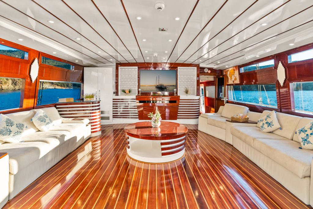A luxurious yacht interior with polished wood floors, cream-colored sofas, decorative pillows, a round coffee table, large windows with water views, and elegant white-and-wood wall accents.