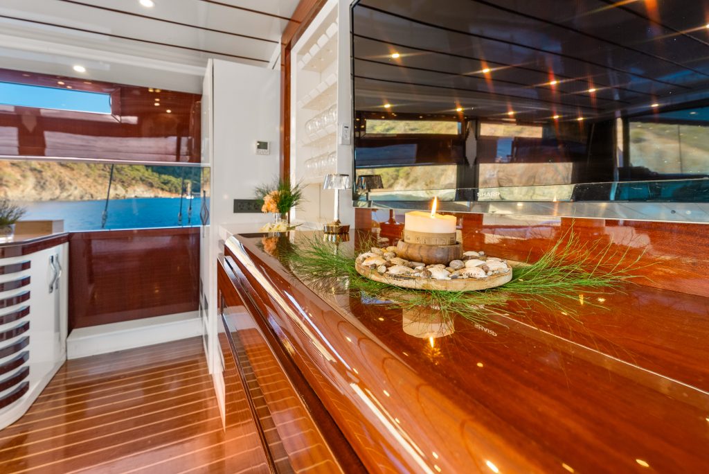 A polished wooden counter inside a yacht, decorated with a lit candle surrounded by pebbles and greenery, with large windows overlooking blue water and a scenic coastline in the background.