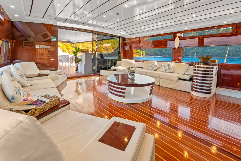 Spacious yacht interior with glossy wooden floors, cream sofas, a round coffee table, and large windows offering views of water and trees outside. Daylight fills the luxurious lounge.