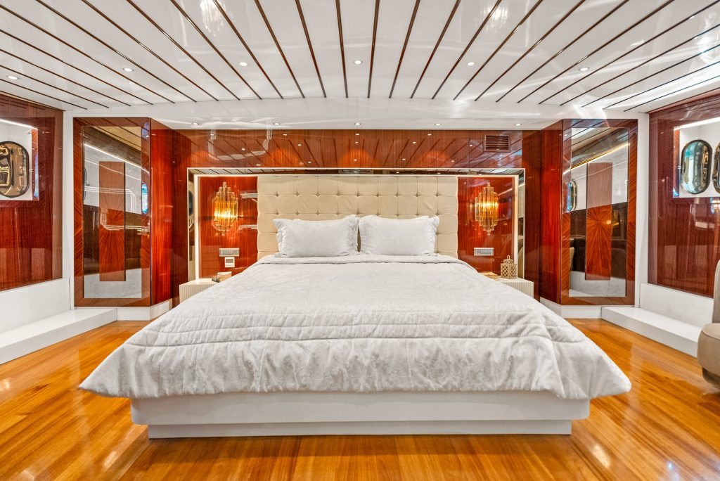 A luxurious bedroom with a large bed featuring white bedding, a tufted headboard, glossy wood paneling, modern lighting, and polished wood floors, creating an elegant and contemporary atmosphere.