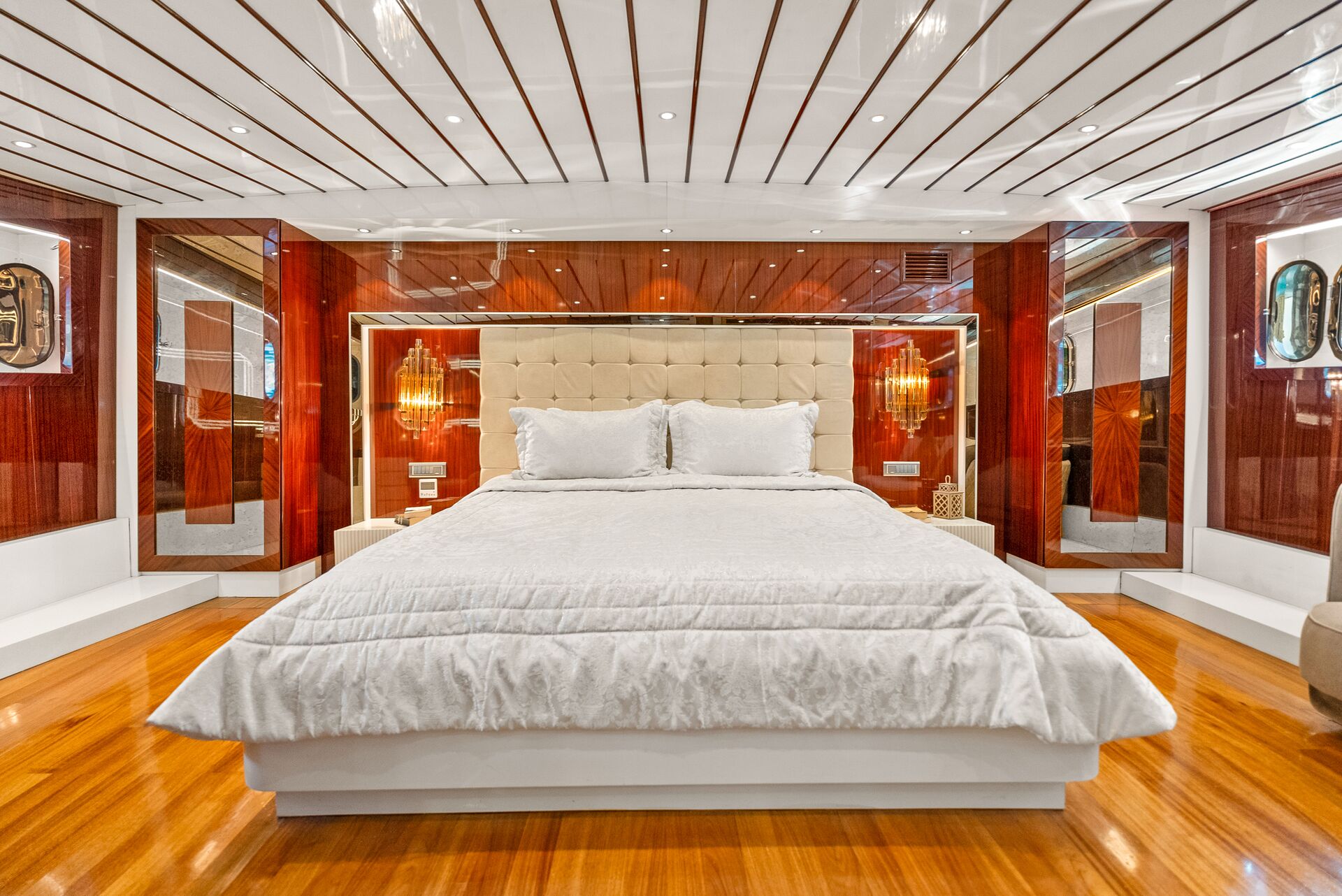 A luxurious bedroom with a large bed featuring white bedding, a tufted headboard, glossy wood paneling, modern lighting, and polished wood floors, creating an elegant and contemporary atmosphere.