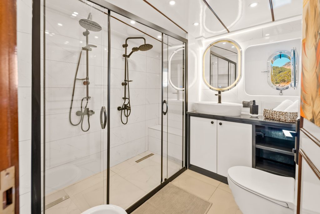 Modern bathroom with double glass shower, two rainfall showerheads, white tile walls, round illuminated mirror above a vessel sink, black countertop, white cabinets, toilet, and a small window showing a scenic outdoor view.