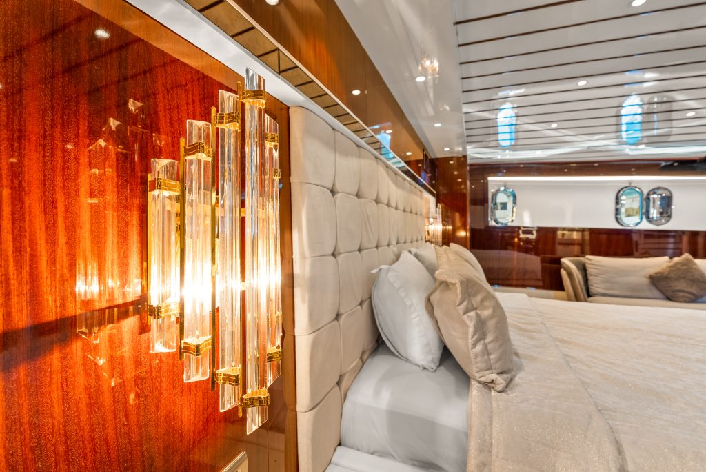 Close-up of a luxurious bedroom on a yacht, featuring a tufted headboard, gold and crystal wall lamp, plush bedding, and wood-paneled walls with porthole windows in the background.