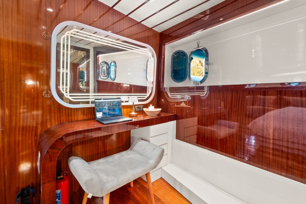 A modern yacht cabin with glossy wood walls, a built-in desk holding a laptop and bowl, a padded gray stool, white cabinetry, and two small windows letting in natural light.