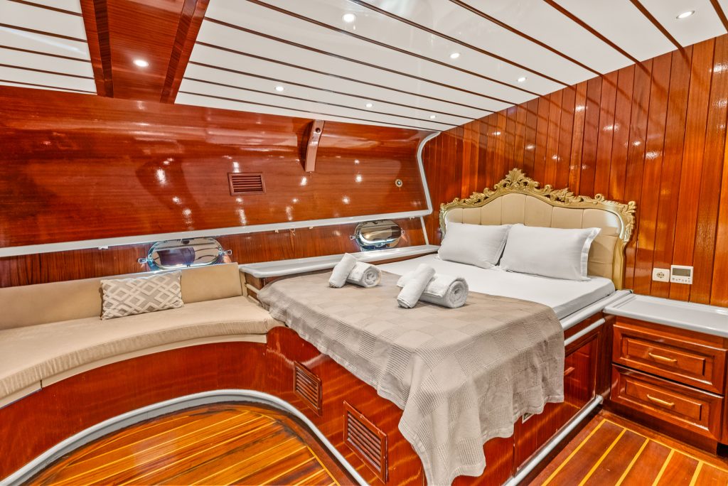 Luxurious yacht cabin with polished wood walls and ceiling, a large bed with white linens and rolled towels, ornate headboard, and a cushioned seating area under small round windows.