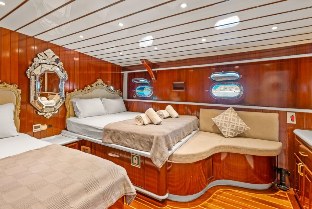 Luxurious yacht cabin with polished wood walls and ceiling, two beds with beige and white bedding, a cushioned bench, elegant mirror, small windows, and soft ambient lighting. Rolled towels are neatly placed on one bed.