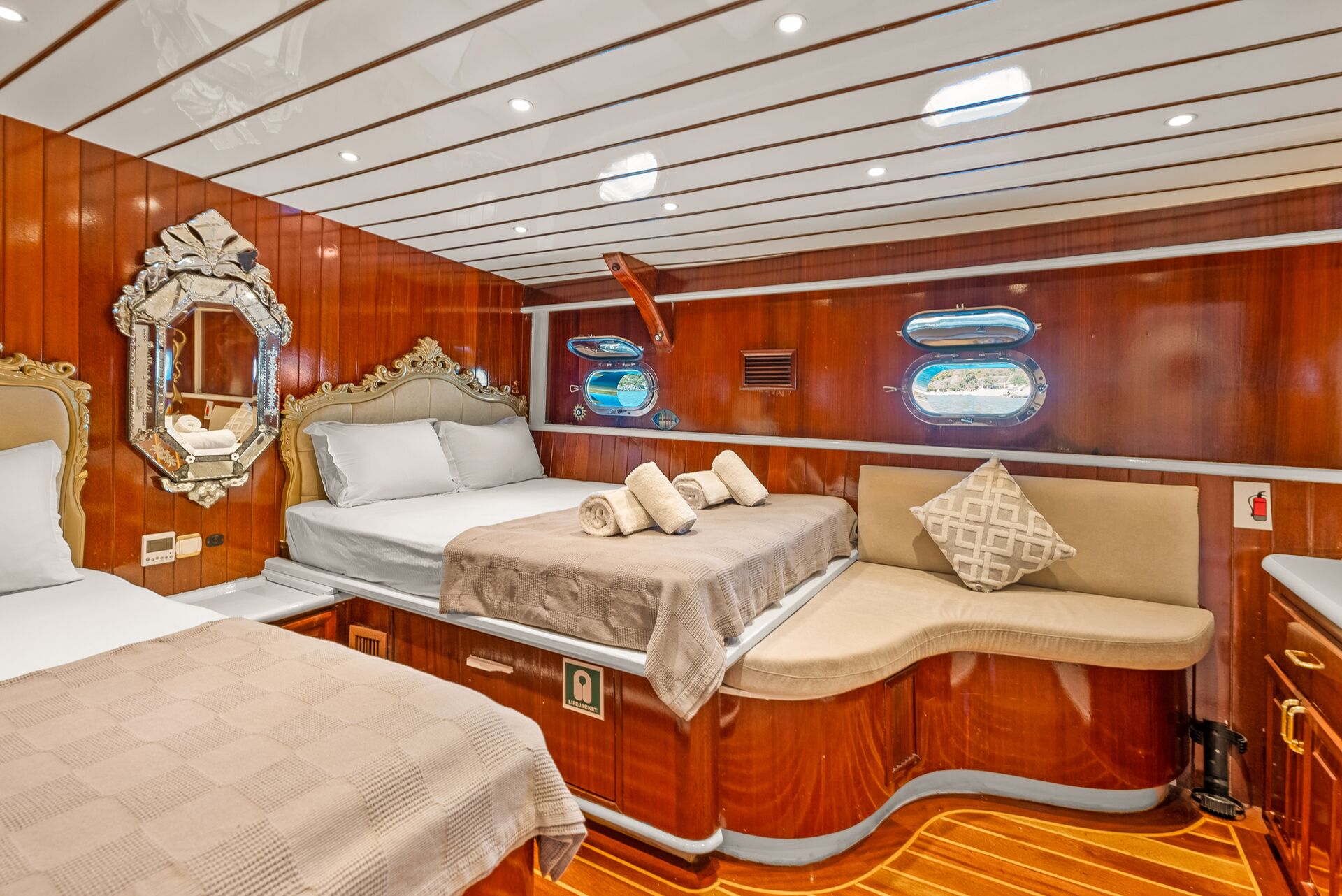 Luxurious yacht cabin with polished wood walls and ceiling, two beds with beige and white bedding, a cushioned bench, elegant mirror, small windows, and soft ambient lighting. Rolled towels are neatly placed on one bed.