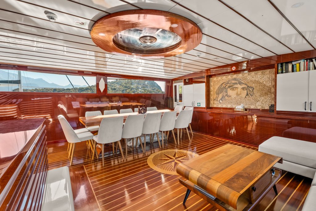 Luxurious yacht interior with polished wood floors, a long dining table surrounded by white chairs, a stylish ceiling fixture, built-in shelves, wall art featuring a classical face, and large windows overlooking mountains.