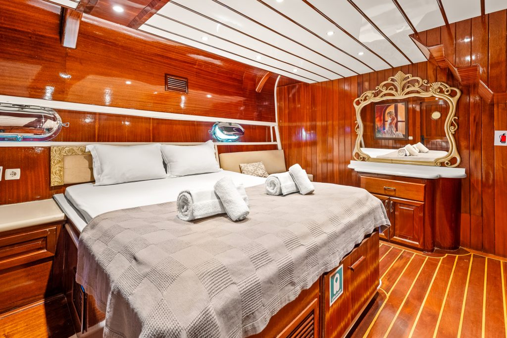 A luxurious wooden cabin with a large bed, white pillows, and rolled towels. The room features glossy wood paneling, a decorative mirror, a painting, and warm lighting, creating a cozy and elegant atmosphere.