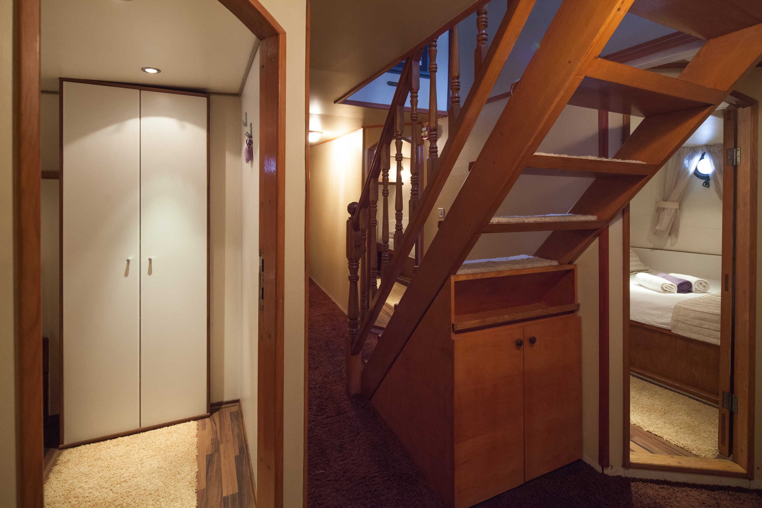 ROMANCA A cozy interior with wooden stairs, a small closet, brown carpet, and a glimpse into a warmly lit bedroom with a bed, towels, and wall-mounted reading light.