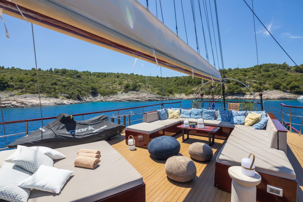 ROMANCA A luxurious yacht deck with cushioned seating, pillows, towels, and poufs overlooks clear blue water and a green, tree-covered shoreline under a sunny sky. A jet ski is also visible on the deck.