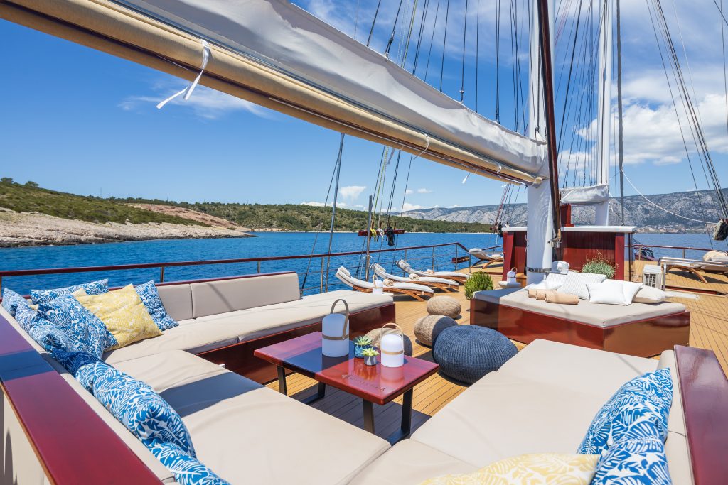 ROMANCA Luxury yacht deck with beige cushioned seating, blue and yellow patterned pillows, and a wooden table, set against clear blue water and a rocky coastline under a partly cloudy sky.