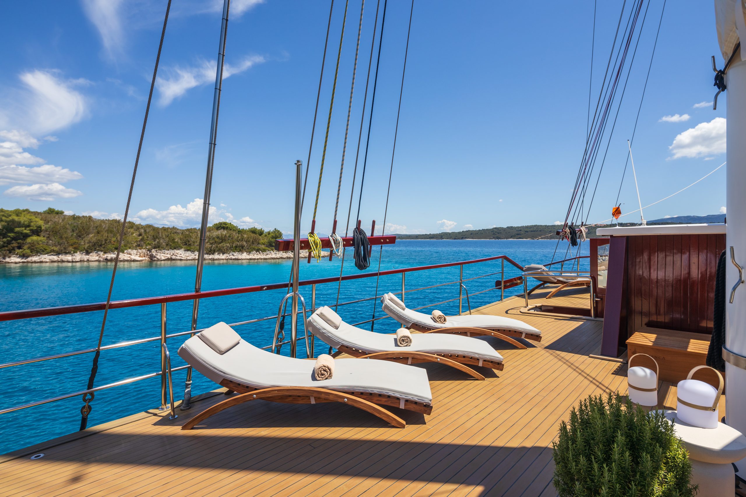 ROMANCA Four cushioned lounge chairs with rolled towels sit on the wooden deck of a boat, overlooking calm blue water and a tree-lined shore under a bright, sunny sky.