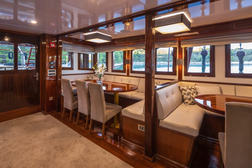 ROMANCA A luxurious yacht dining area with polished wood interior, cushioned bench seating around glossy round tables, elegant chairs, a flower arrangement, and large windows showing water and greenery outside.