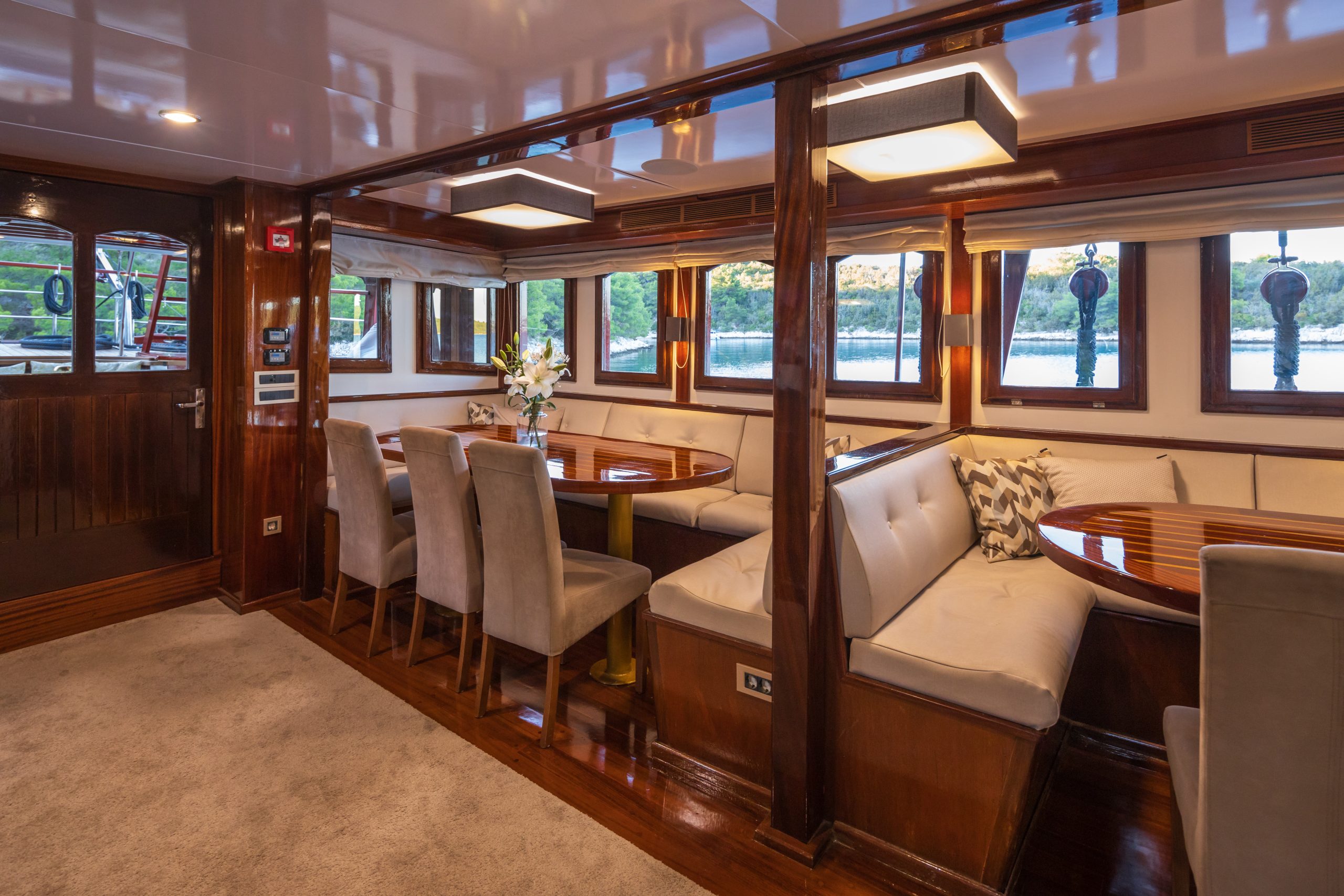 ROMANCA A luxurious yacht dining area with polished wood interior, cushioned bench seating around glossy round tables, elegant chairs, a flower arrangement, and large windows showing water and greenery outside.