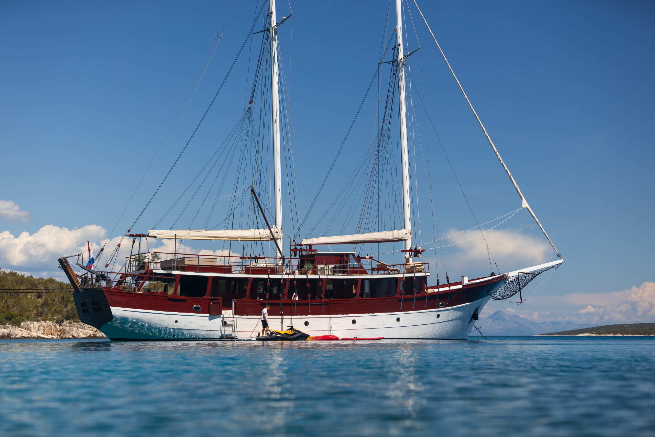 ROMANCA A large, classic wooden sailing yacht with two masts is anchored in calm blue water near a rocky shoreline under a clear sky. Kayaks float beside the boat.