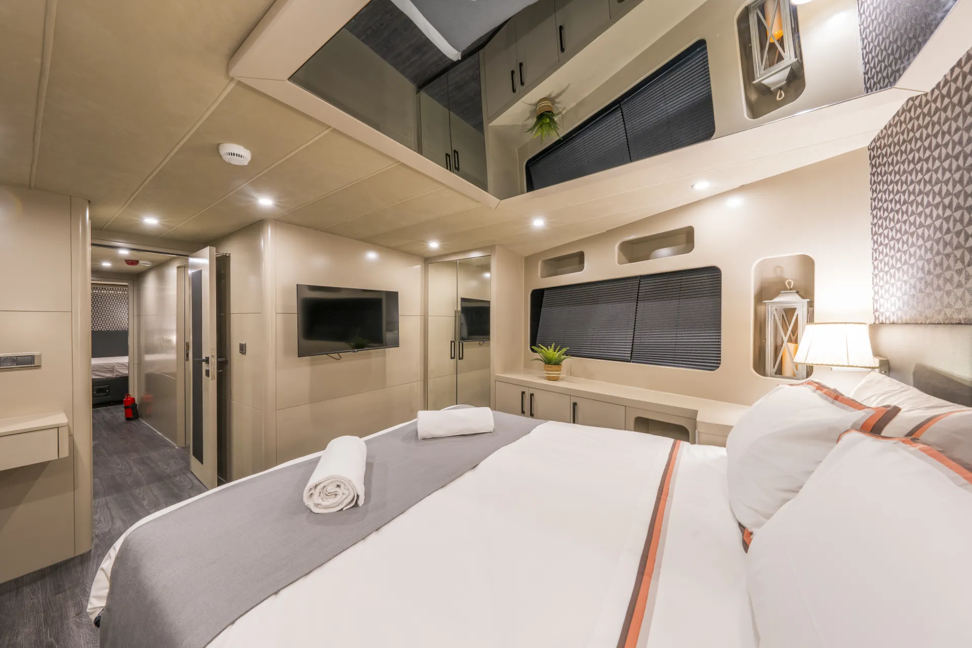JUNIOR SIMAY A modern RV bedroom with a large bed, neatly rolled towels, wall-mounted TV, mirrored cabinets, windows with blinds, soft lighting, and neutral-toned decor.