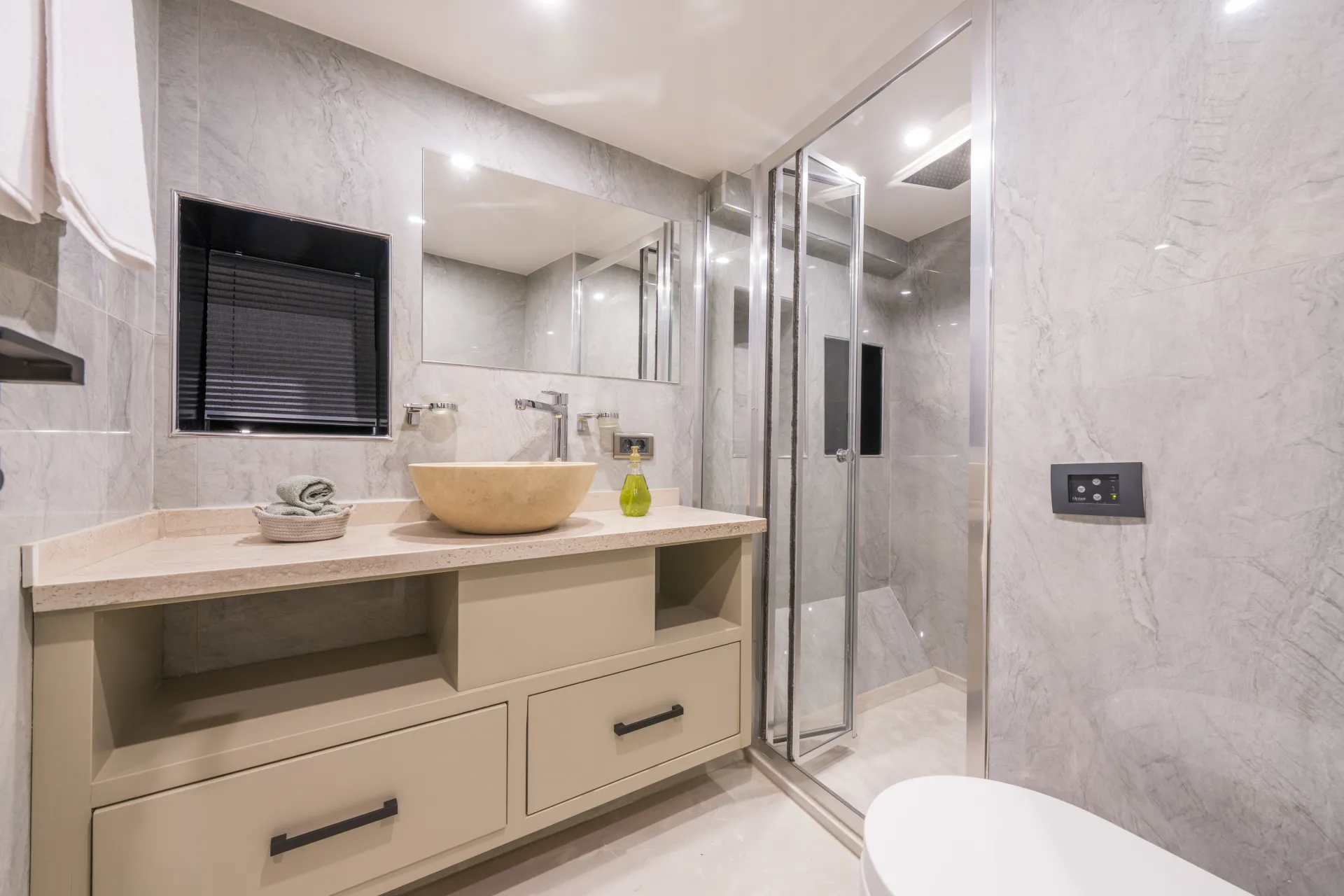 JUNIOR SIMAY Modern bathroom with light beige marble walls, a walk-in glass shower, a large mirror, a vessel sink on a beige countertop with drawers below, and a round toilet in the foreground. Towel and soap are also visible.