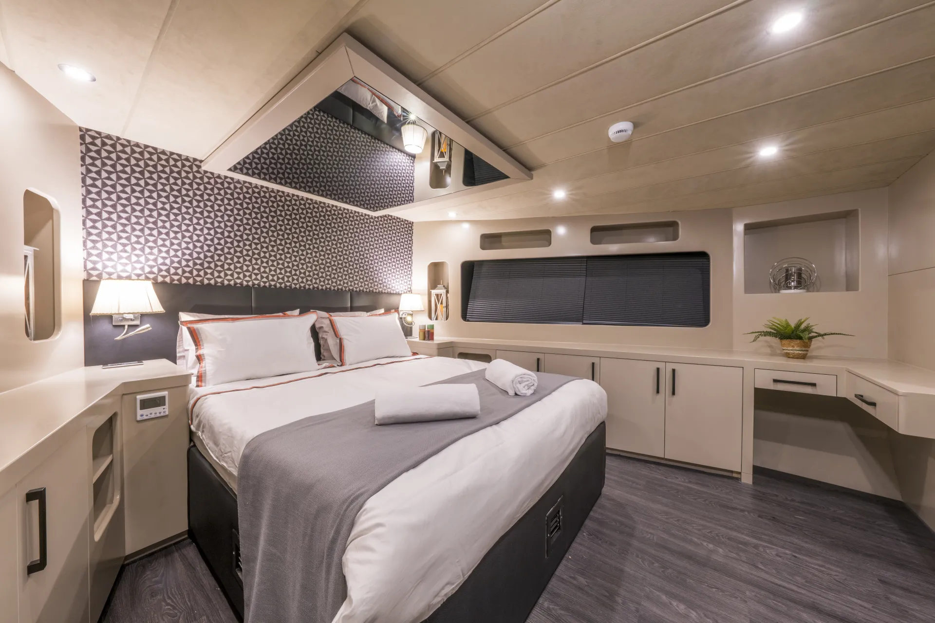 JUNIOR SIMAY A modern, well-lit bedroom with a large bed covered in white and gray linens, two folded towels, geometric accent wall, built-in shelves, a desk, overhead lighting, and a large window with a dark shade.