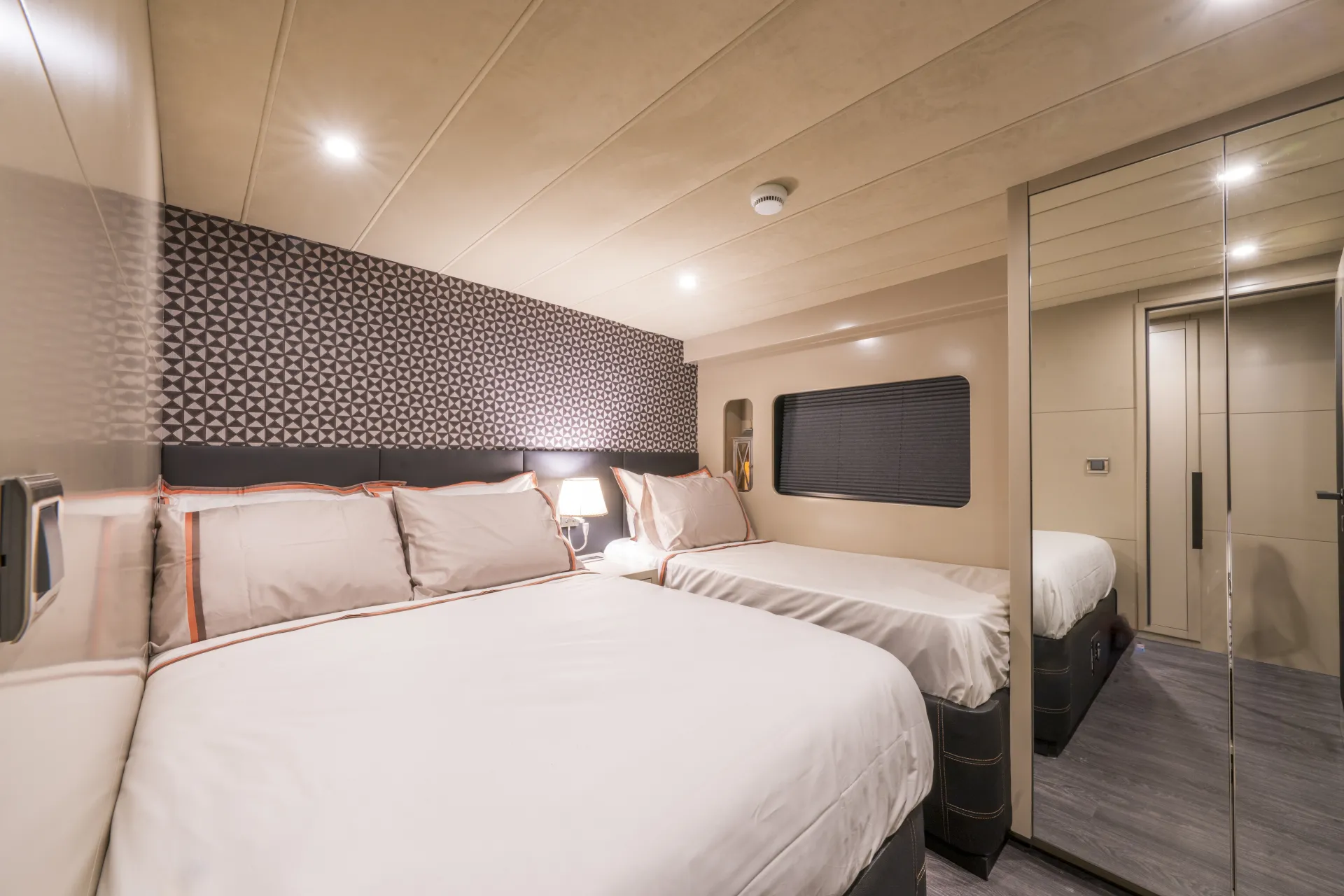 JUNIOR SIMAY Modern RV bedroom with two large beds, patterned accent wall, a nightstand with a lamp, window with a shade, mirrored closets, and soft recessed lighting, creating a cozy and spacious atmosphere.