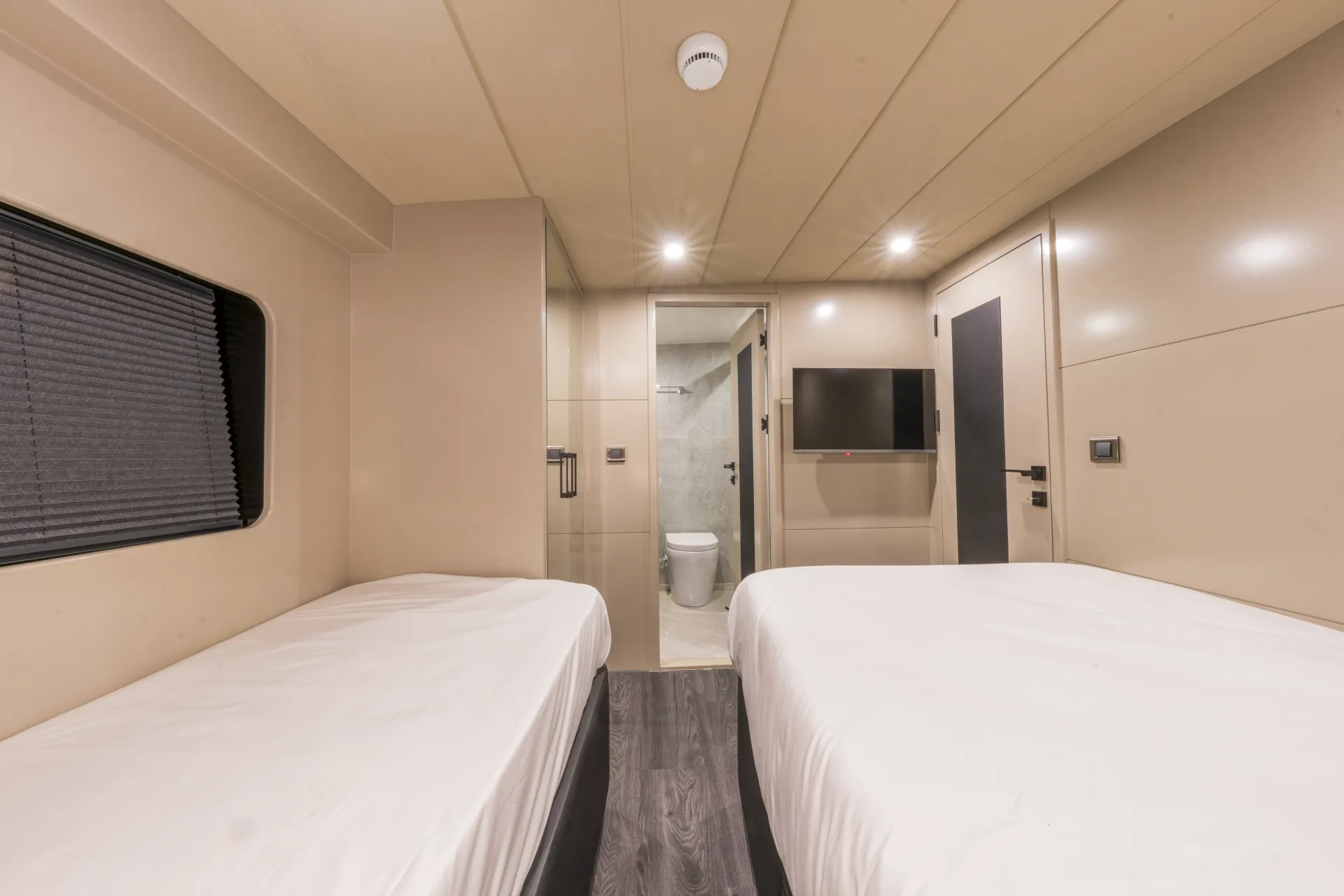 JUNIOR SIMAY A modern, minimalistic room with two single beds, beige walls, a small wall-mounted TV, and a window with a closed blind. There is a doorway leading to a bathroom with a visible toilet.