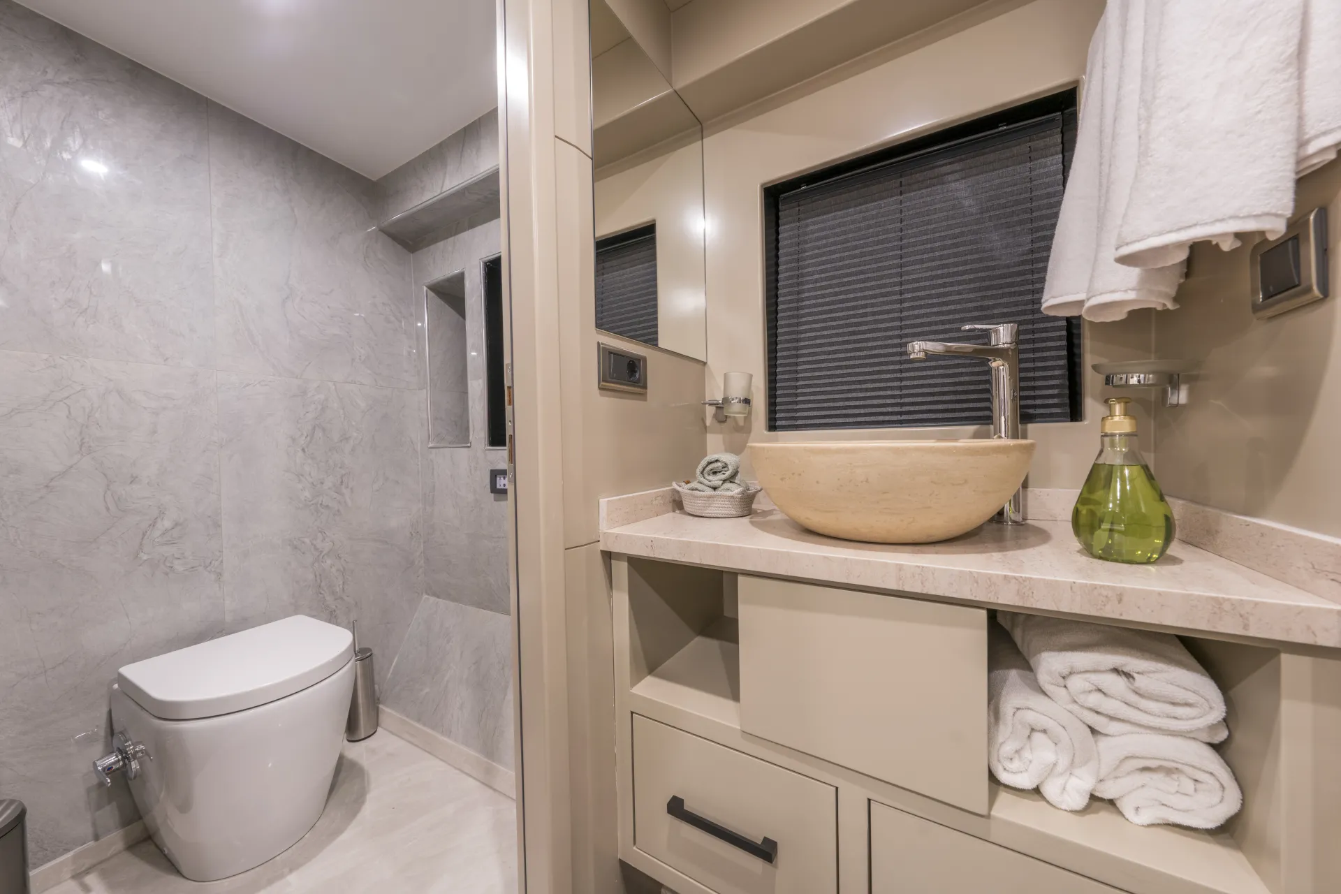 JUNIOR SIMAY Modern bathroom with gray marble walls, a white toilet, a beige vanity with a round stone sink, a tall silver faucet, rolled white towels, and a green soap dispenser; towels hang above the sink.