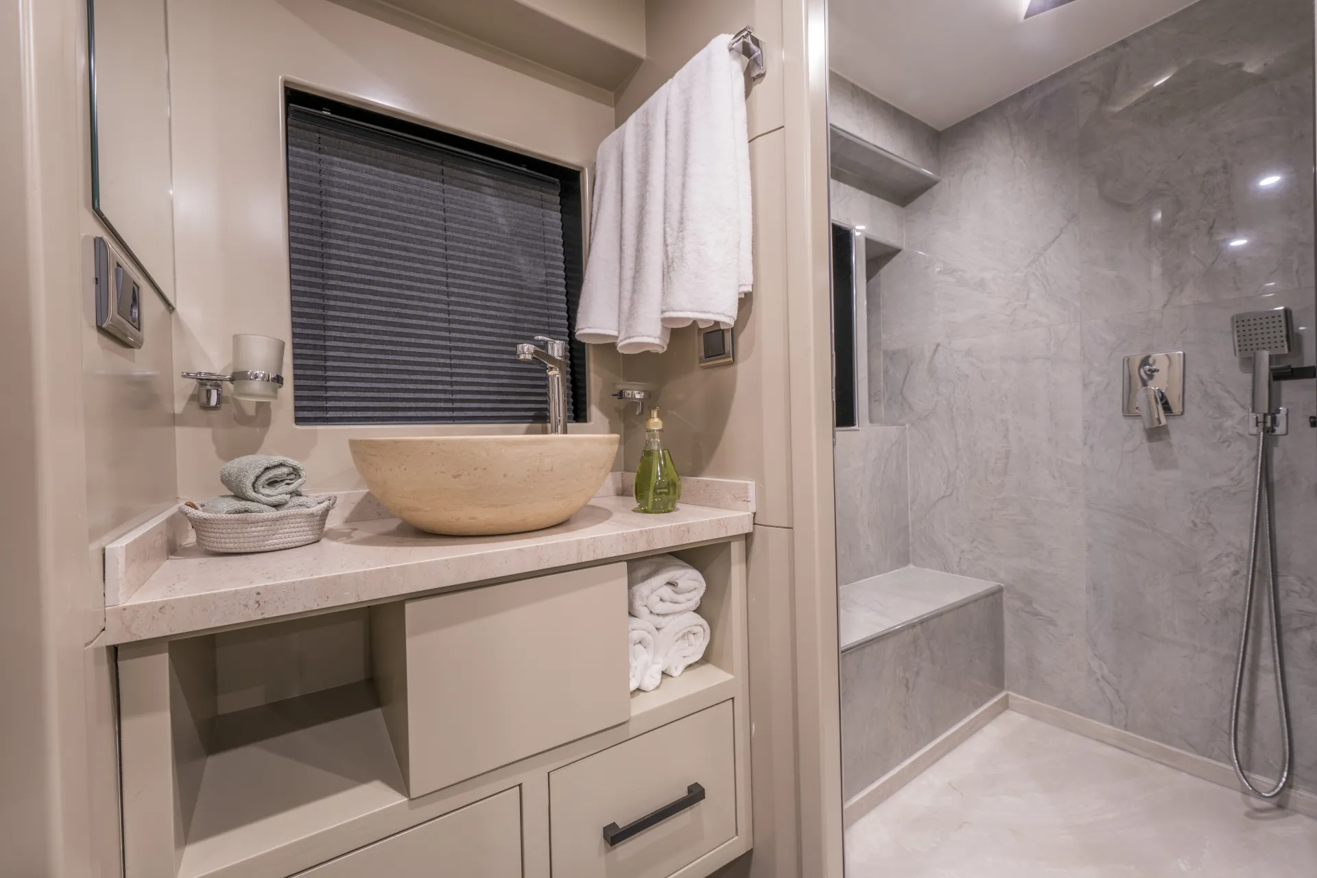 JUNIOR SIMAY Modern bathroom with a stone sink, rolled towels, and a glass soap dispenser on a beige vanity. A towel hangs above, and a walk-in shower with a marble wall and built-in bench is on the right.